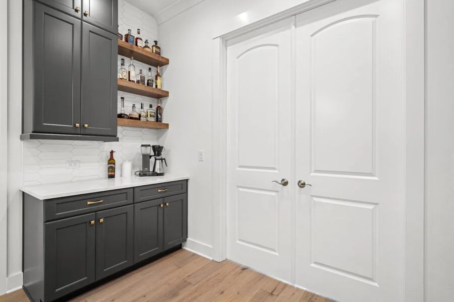 Stylish home bar area with dark gray cabinets, wooden open shelves with liquor bottles, a coffee machine, and white hexagonal backsplash in a modern kitchen setting.