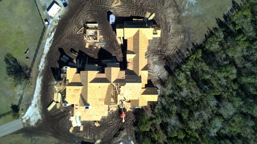 Aerial view of a large, newly constructed residential home with multiple sections, surrounded by trees and construction site.