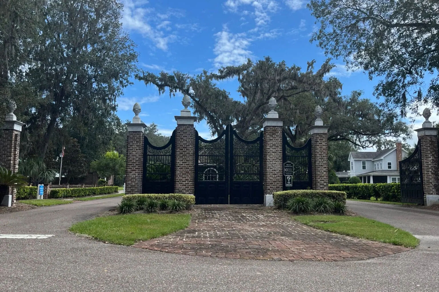 Gated entrance to a luxurious residential community with brick and wrought iron gates, lush landscaping, and large oak trees under a blue sky with scattered clouds, representing upscale home development.