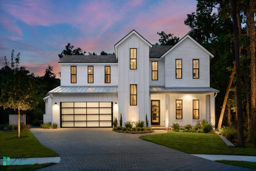 A spacious modern farmhouse with white exterior siding, large windows, and an attached two-car garage, set against a scenic sunset sky with lush landscaping.