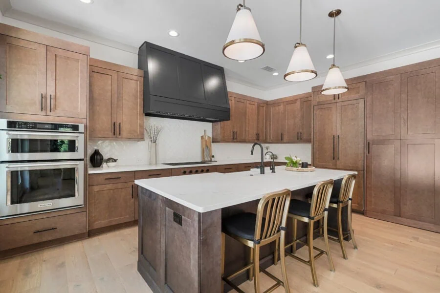 A modern kitchen featuring warm wood cabinets, a large white island with seating, stainless steel appliances, black fixtures, and a stylish black range hood, illuminated by contemporary pendant lights.
