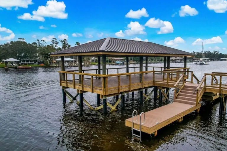 A scenic waterfront dock with a covered pavilion and a small ramp leading into the water, surrounded by calm water, lush trees, and neighboring houses under a bright blue sky with fluffy clouds.