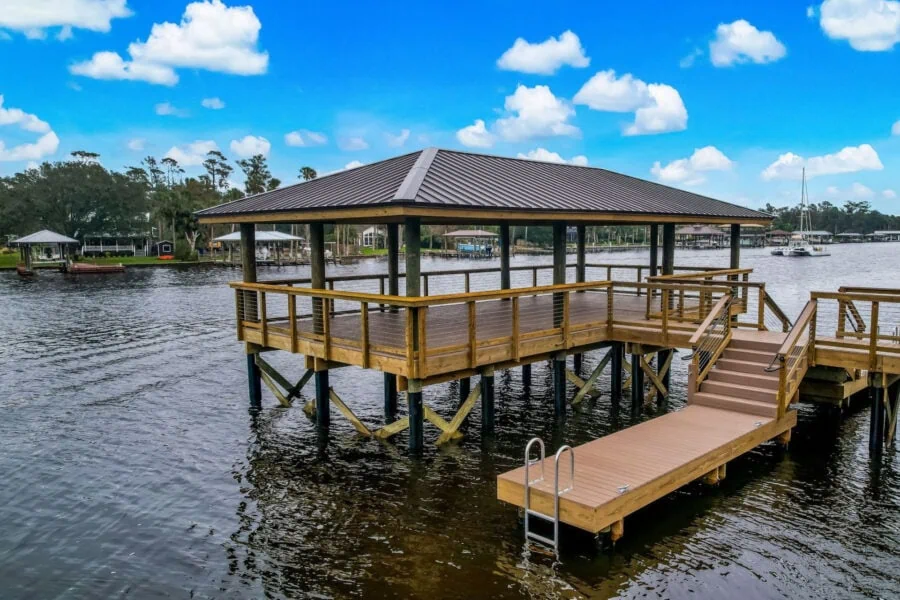 A scenic waterfront dock with a covered pavilion and a small ramp leading into the water, surrounded by calm water, lush trees, and neighboring houses under a bright blue sky with fluffy clouds.