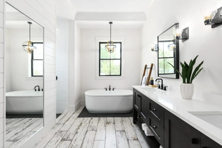 Luxurious modern bathroom featuring a freestanding bathtub, stylish black fixtures, and a sleek black vanity with white countertops illuminated by natural light from dual windows. Elegant wall-mounted lighting and contemporary decor create a spa-like ambiance.