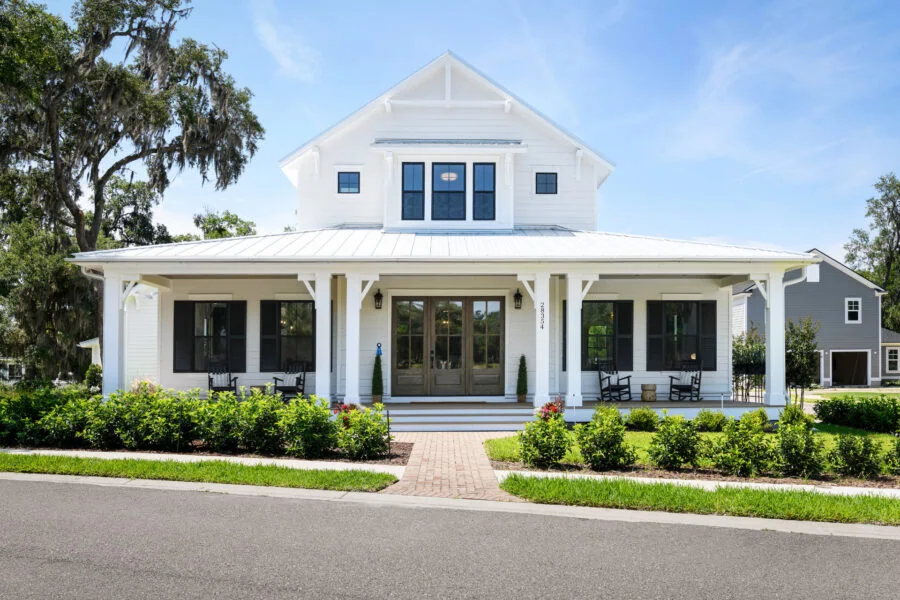 Luxurious White Modern Farmhouse with Covered Front Porch and Black Accents, Surrounded by Beautiful Landscaping and Tall Trees under a Bright Blue Sky.