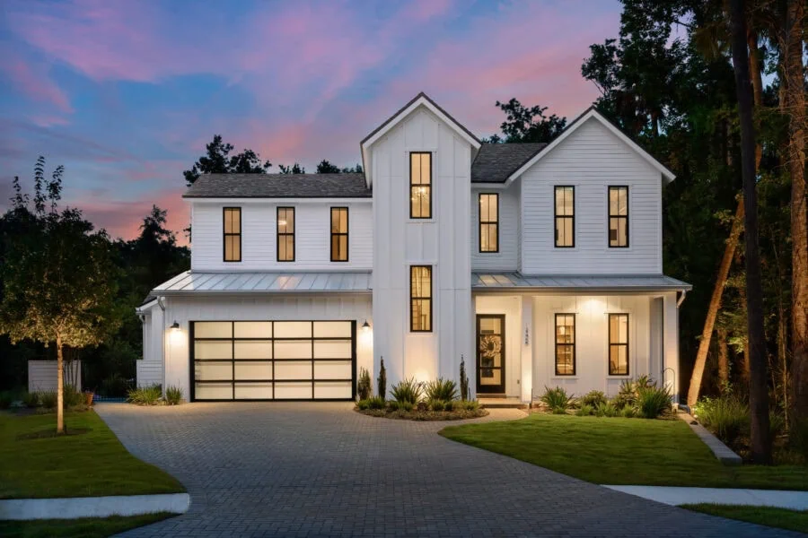 Modern luxury white farmhouse-style home with large windows and a spacious driveway, illuminated at dusk against a colorful sky, showcasing elegant architecture and landscaped yard.