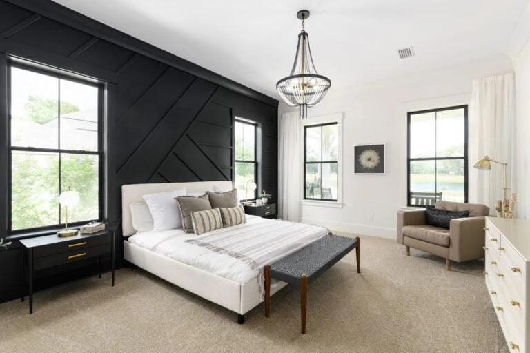 Modern bedroom interior with black accent wall, elegant chandelier, and large windows letting in natural light, featuring a cozy bed, stylish side tables, and a sitting area with a beige armchair.