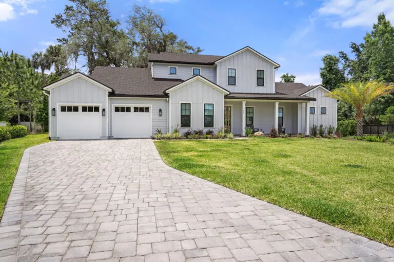 Modern white farmhouse-style home with a spacious driveway, manicured lawn, and lush landscaping, showcasing custom construction quality and curb appeal.