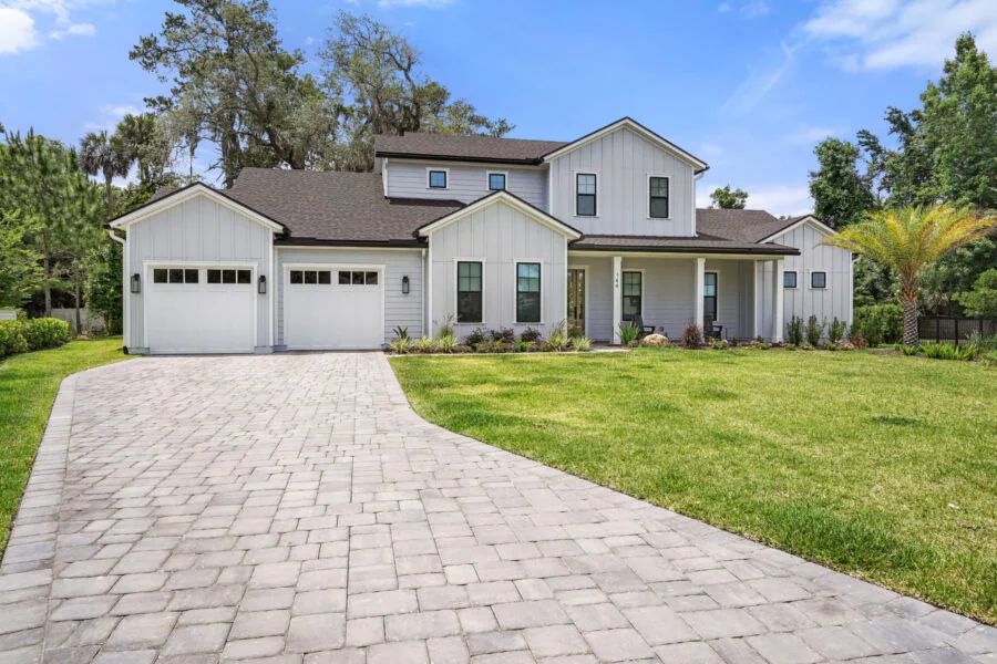 Modern white farmhouse-style home with a spacious driveway, manicured lawn, and lush landscaping, showcasing custom construction quality and curb appeal.