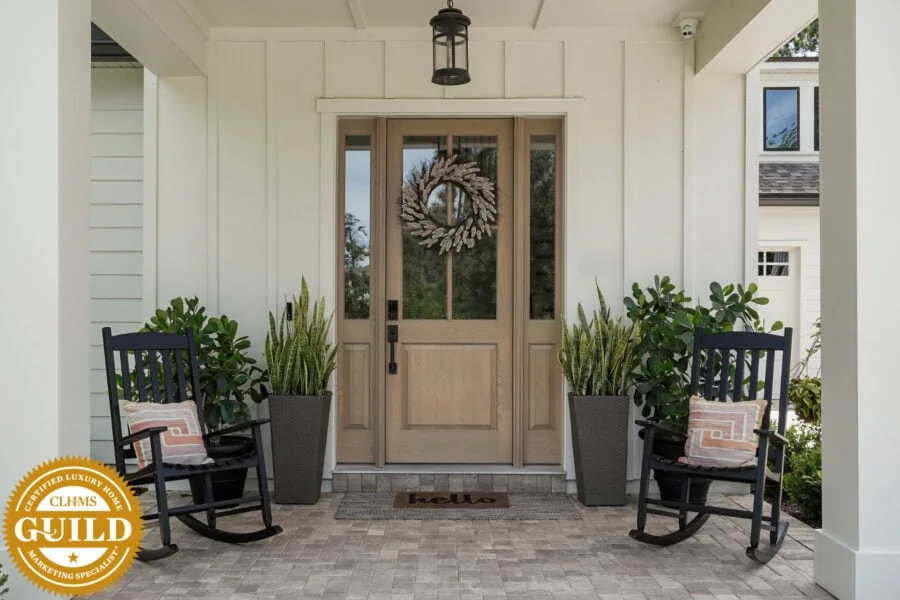 Elegant front porch with a natural wood door decorated with a wreath, flanked by potted greenery, black rocking chairs with pillows, and a welcoming hello doormat, highlighting charming curb appeal and outdoor sophistication.