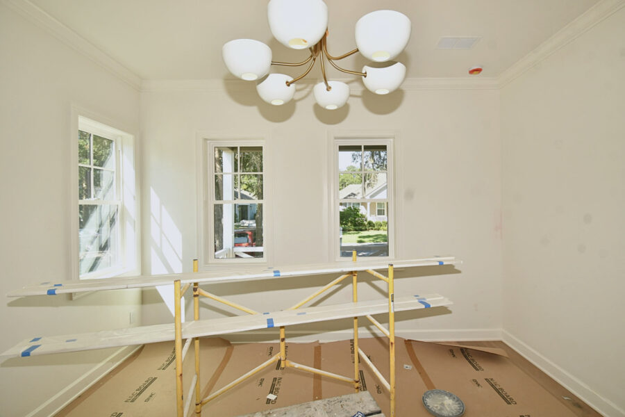 A newly built dining space featuring white walls, three large windows allowing natural light, and a sleek modern chandelier, with construction materials still present, showcasing recent progress in home development.