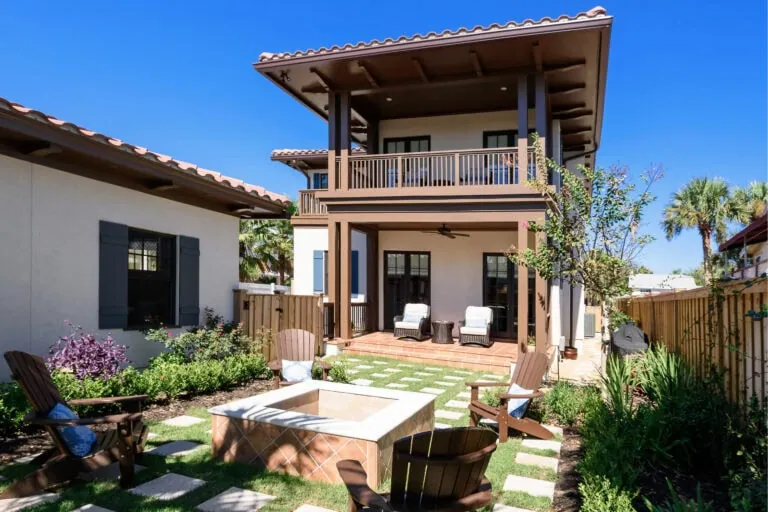 Elegant backyard patio with outdoor seating and fire pit, featuring a two-story house with a spacious balcony, surrounded by lush greenery and a clear blue sky.