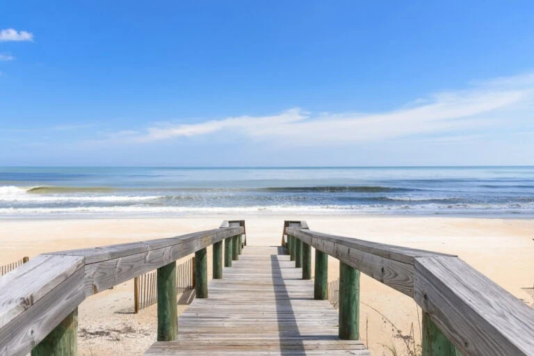 Stress-free beach access pathway leading to the ocean with clear blue skies, gentle waves, and soft sandy shores, perfect for relaxation, leisurely walks, and enjoying coastal scenery.