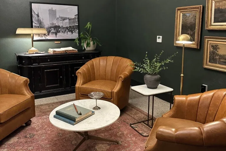 Cozy live/work room with vintage leather armchairs, a marble coffee table, and classic artwork on dark green walls, creating a warm and inviting atmosphere perfect for relaxation or conversation.