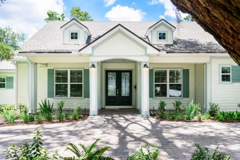 Charming modern farmhouse-style home with mint green shutters, a welcoming covered porch, and a beautifully landscaped front yard under a bright, sunny sky.