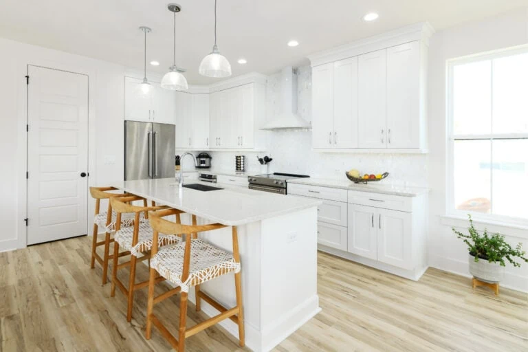 Bright and modern white kitchen with a large island, wooden chairs, stainless steel appliances, and ample natural light from a large window, showcasing stylish cabinetry and a clean, minimalist design.