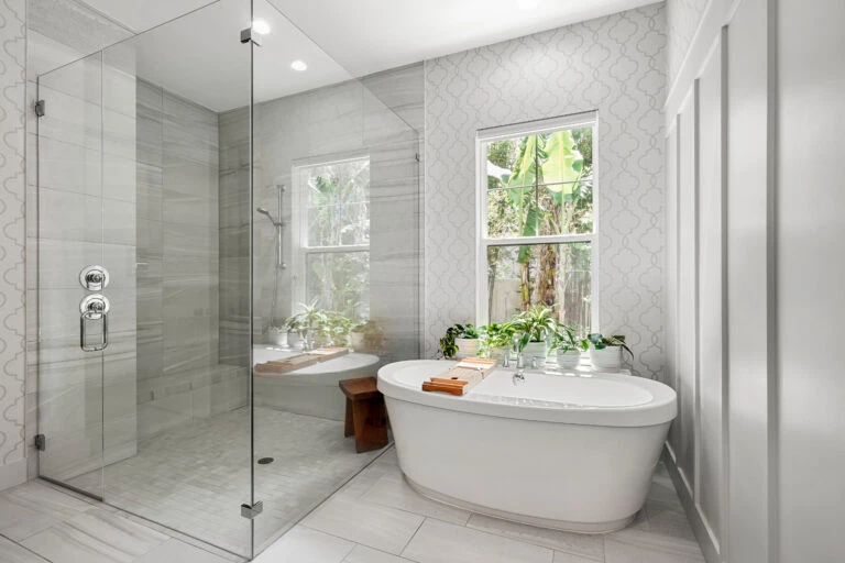 Luxurious bathroom with a walk-in glass shower, freestanding bathtub, and natural light from large windows overlooking greenery. Elegant, modern design with white and neutral tones, perfect for relaxation and self-care in a beautifully crafted home.