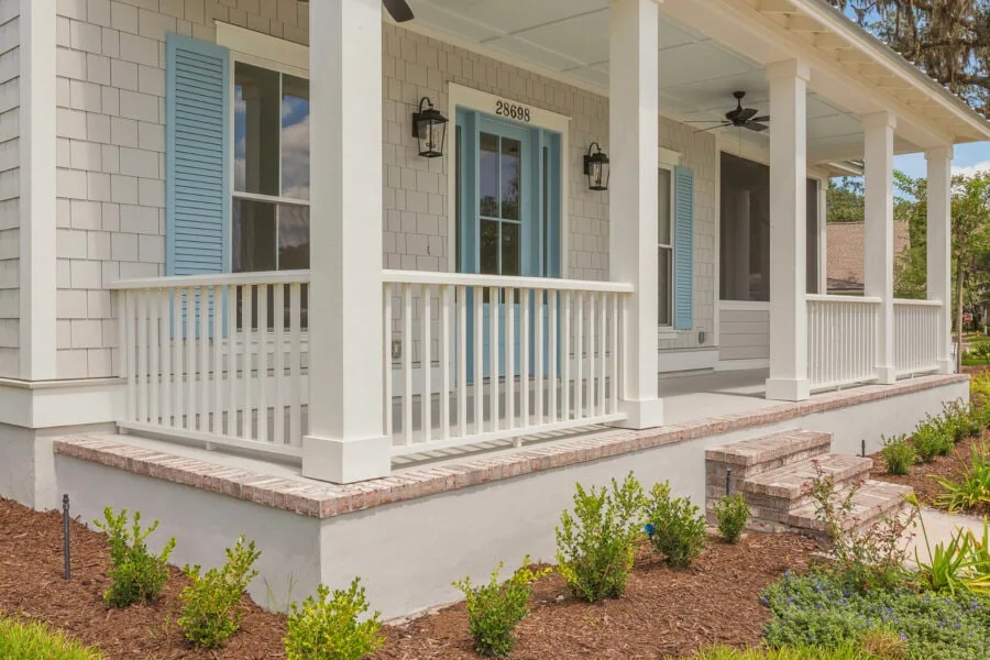 Front porch of a new home showcasing a welcoming entryway with white railings, brick steps, and light-colored exterior siding accented by blue shutters, surrounded by landscaped greenery.