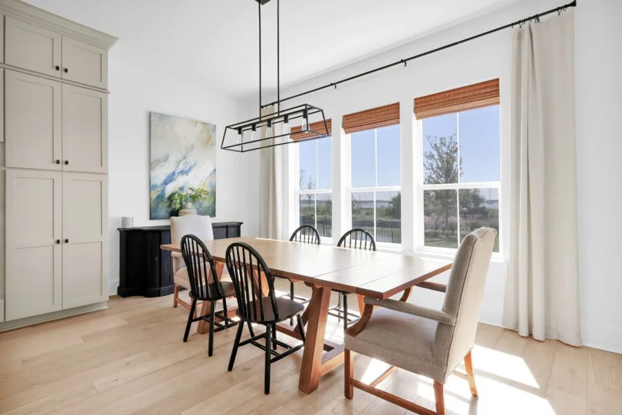 Elegant dining room featuring a wooden table accompanied by black and beige chairs, large windows with natural light, white walls, and a modern black chandelier, creating a bright and inviting space for family gatherings and meals.
