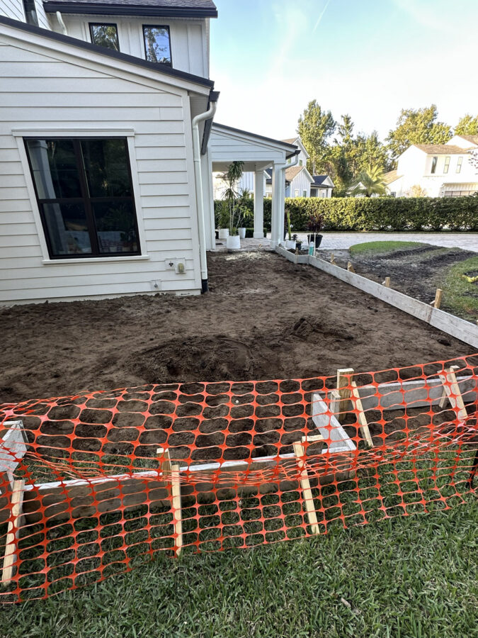 New construction home foundation preparation with dirt work in progress, white house siding, and landscaping underway at a residential property.