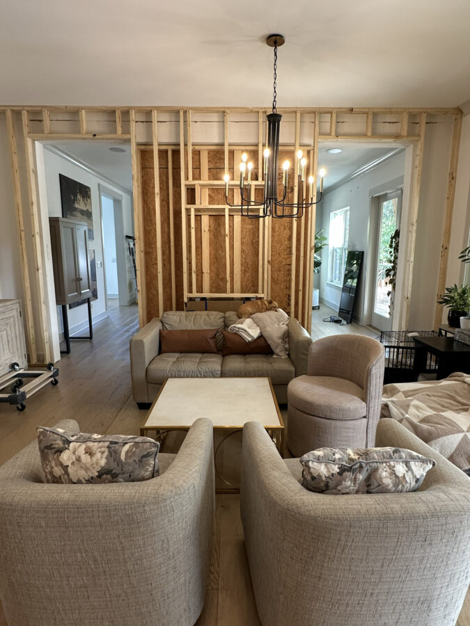 Modern living room with unfinished wall framing, featuring a beige sofa, floral armchairs, a round side chair, a square coffee table, and a black chandelier, highlighting a home renovation or construction project.
