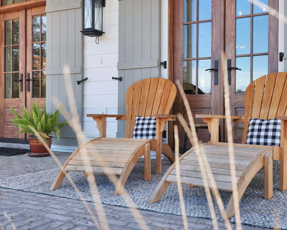 Adirondack wooden chairs with black and white checkered pillows on a porch with a textured rug and potted greenery.