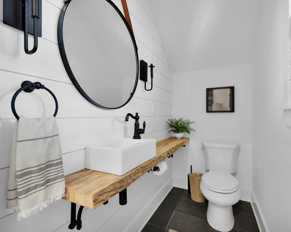Contemporary bathroom with a rustic wood vanity, black fixtures, a round mirror, and modern decor, featuring white shiplap walls, dark floor tiles, and a small window with shutters.