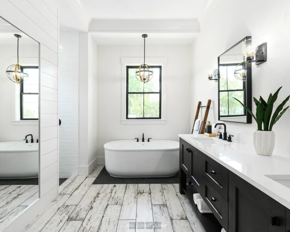 Bright modern bathroom with a freestanding bathtub, black fixtures, and a double vanity with black cabinets, white countertops, and decorative lighting, featuring natural light from windows.