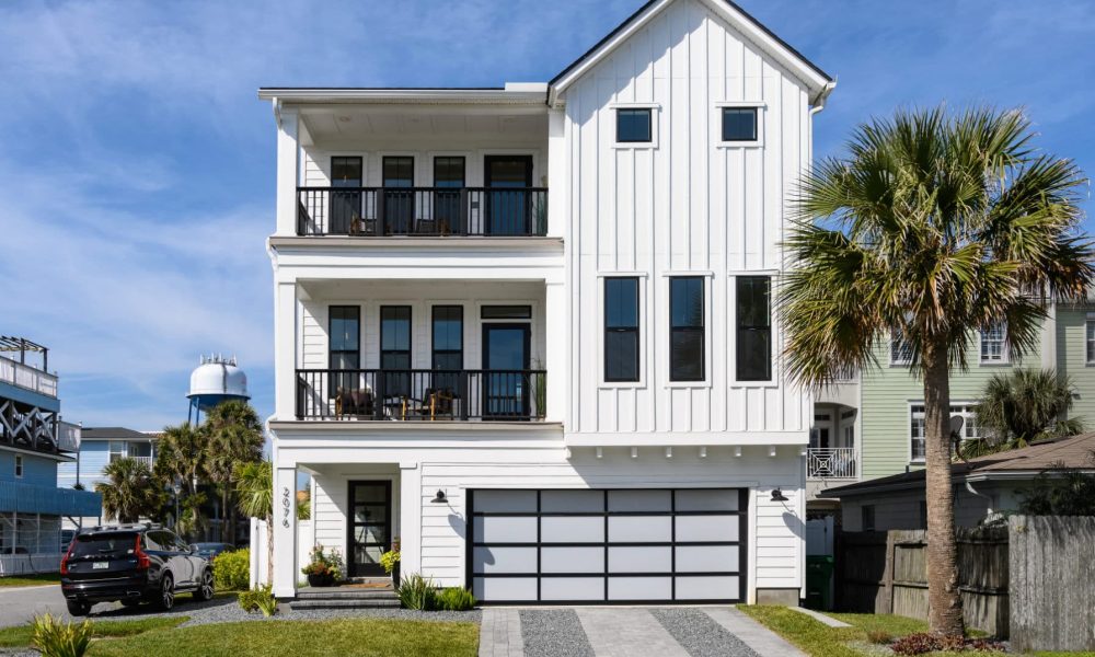 Modern white multi-story coastal home with black accents, large windows, and balconies, featuring a landscaped front yard with a palm tree and a paved driveway, under a bright blue sky.