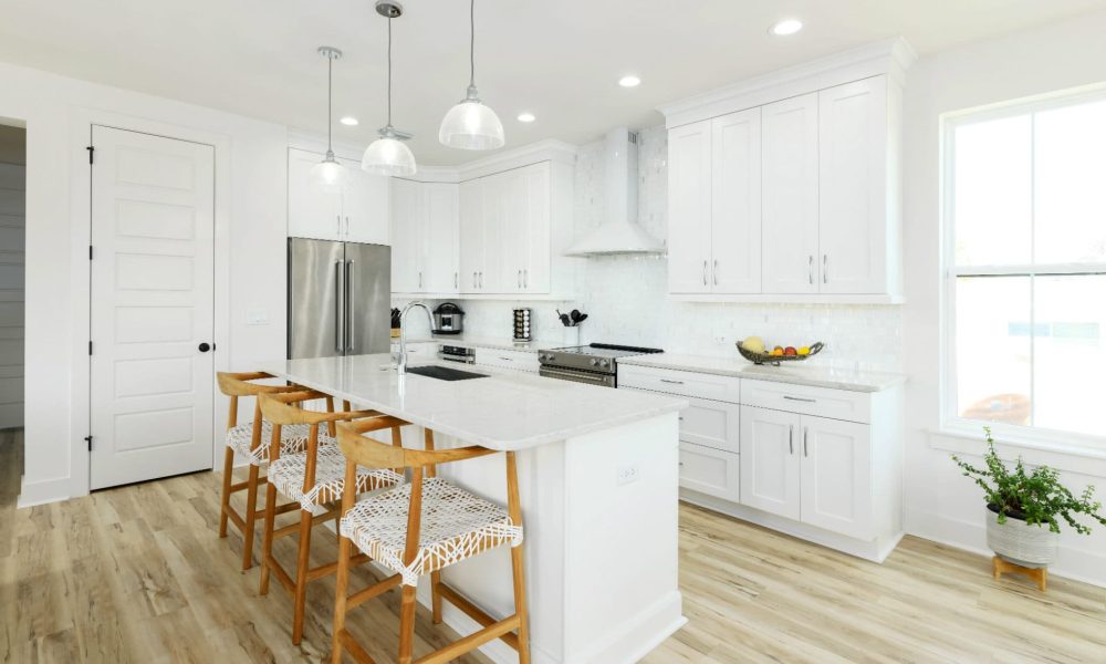 Bright white modern kitchen with cabinetry, stainless steel appliances, and an island with wooden bar stools, featuring natural light from a large window and a light wood floor.