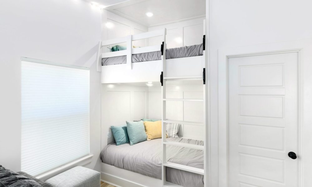 Bright, modern bedroom featuring a built-in bunk bed with a lower full-size bed and an upper twin bed, all finished in white with cozy bedding and decorative pillows; natural light streaming through a large window.