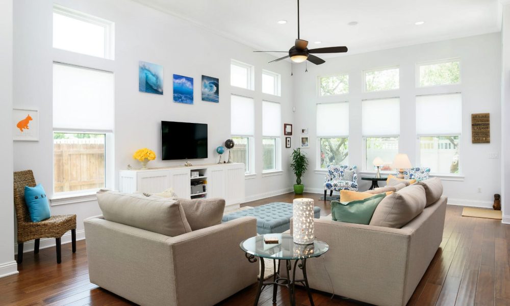 Bright and spacious living room with white walls, high ceilings, large windows with blinds, a modern ceiling fan, and stylish furniture and decor for a welcoming atmosphere.