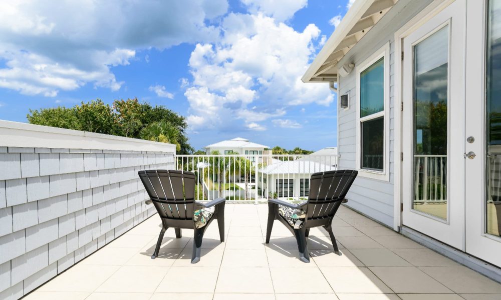 Modern white balcony with outdoor chairs, overlooking a vibrant neighborhood with greenery and a bright blue sky with scattered clouds.