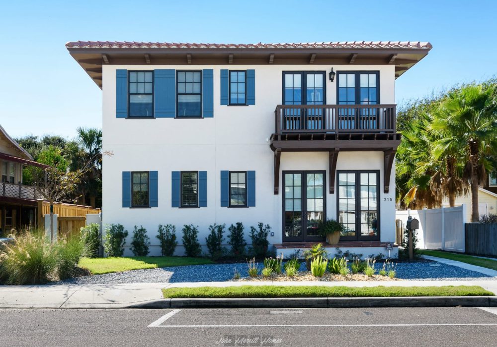 Modern coastal style house featuring white stucco exterior, dark blue window shutters, and a second-story balcony with wooden railing, surrounded by lush landscaping and palm trees.