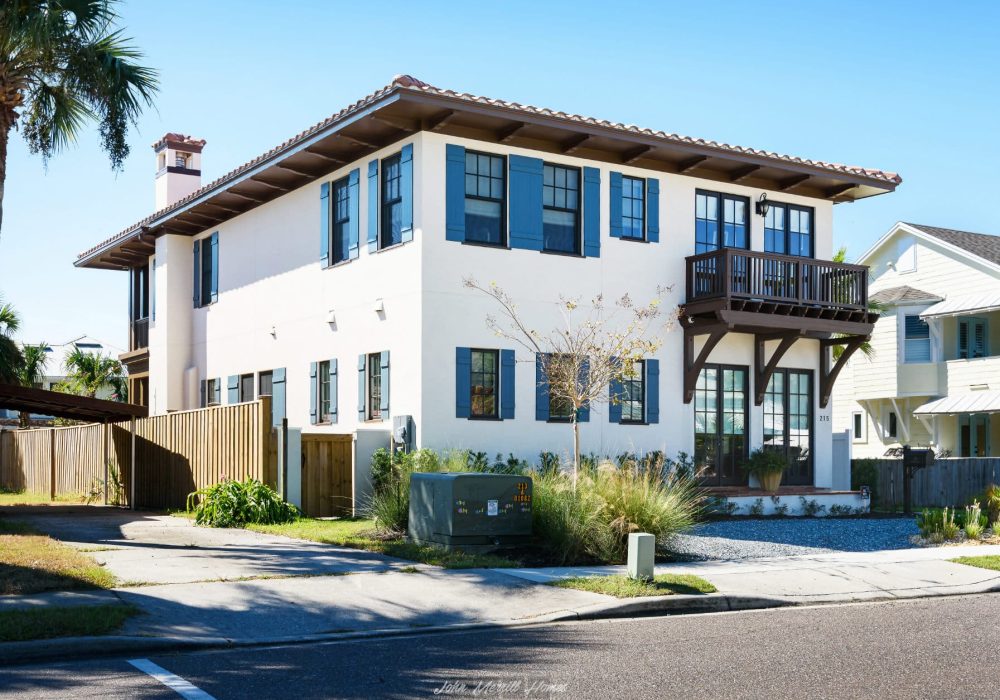 Modern white two-story home with blue window shutters, a small balcony, and landscaped yard in a sunny suburban neighborhood.