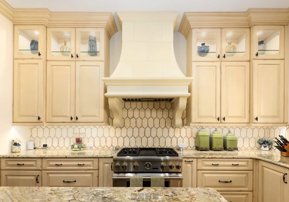 Warm cream-colored kitchen cabinets with black hardware, a matching beige tile backsplash, and a stainless steel stove with a decorative vent hood, creating a cozy and elegant cooking space.