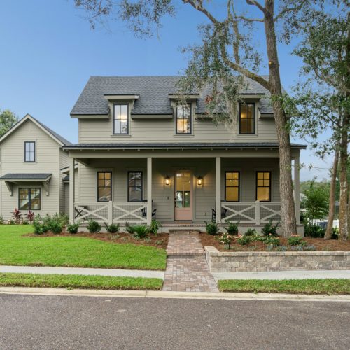 Luxurious modern two-story house with a spacious front porch, surrounded by lush greenery and landscaped garden, showcasing timeless design by John Merrill Homes.