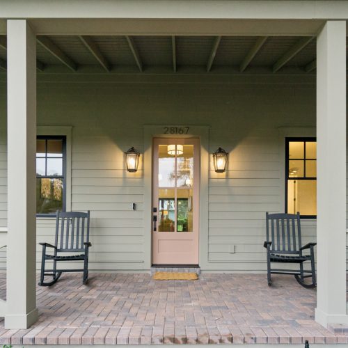 Elegant front porch of a modern house with two black rocking chairs, a pink front door, outdoor lighting fixtures, and a cozy welcoming atmosphere, showcasing home design by John Merrill Homes.