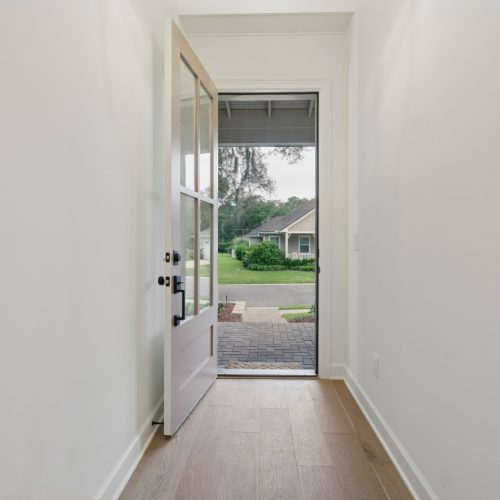 Bright, modern entryway with white walls, light wood flooring, and a glass-paneled door opening to a landscaped front yard and neighborhood.