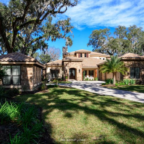Luxury home with stone accents and a lush front yard, surrounded by mature trees under a bright blue sky, showcasing elegant landscaping and a welcoming entrance.