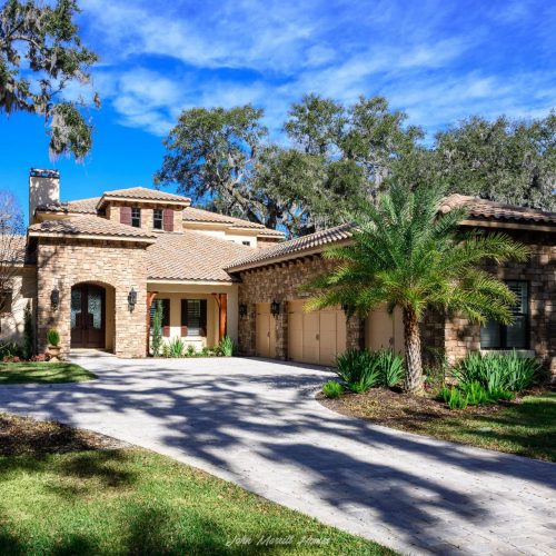Luxurious single-family home with a stone facade, lush landscaping, and a paved driveway under a bright blue sky, showcasing elegant architecture and vibrant greenery.