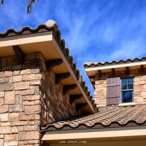 Handcrafted stone and stucco exterior of a luxury home with terracotta roof tiles, sky and clouds in the background.