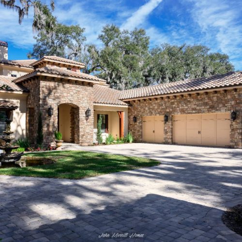 Luxurious single-story home with a stone and stucco exterior, three-car garage, Mediterranean-style clay tile roof, lush landscaping, and a picturesque fountain in the front yard.