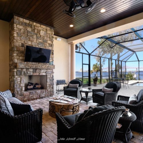 Modern enclosed patio with outdoor seating, stone fireplace, flat-screen TV, and view of water and bridge through screened windows.
