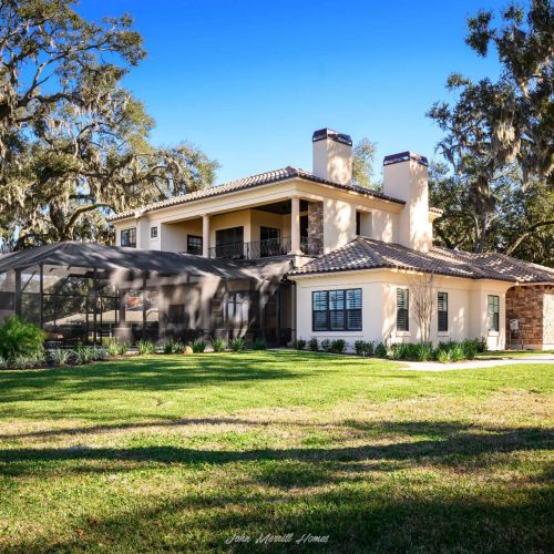 Luxury modern two-story home with a spacious backyard, covered patio, and lush landscaping, surrounded by mature moss-draped trees, under a clear blue sky.