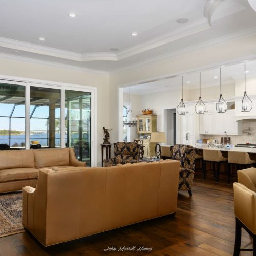 Modern living room with large glass sliding doors offering waterfront views, featuring tan leather sofas, patterned armchairs, and elegant decor, seamlessly connected to a bright, open kitchen and dining area.