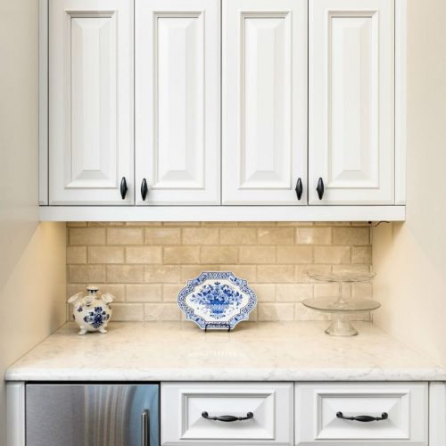 A kitchen cabinet with white paneled doors, black handles, and a beige brick backsplash, featuring decorative blue and white porcelain items on a marble countertop.