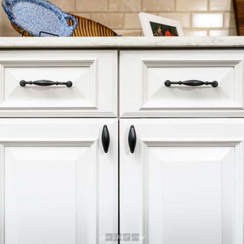 White kitchen cabinets with black handles and raised panel design, granite countertop, decorative items, and framed photo on beige brick wall background.