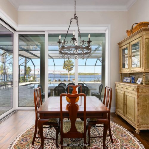 Elegant dining area with a wooden table, vintage chandelier, and a distressed yellow hutch overlooking a screened-in patio with water views. Bright, airy space blending rustic charm with modern comfort.
