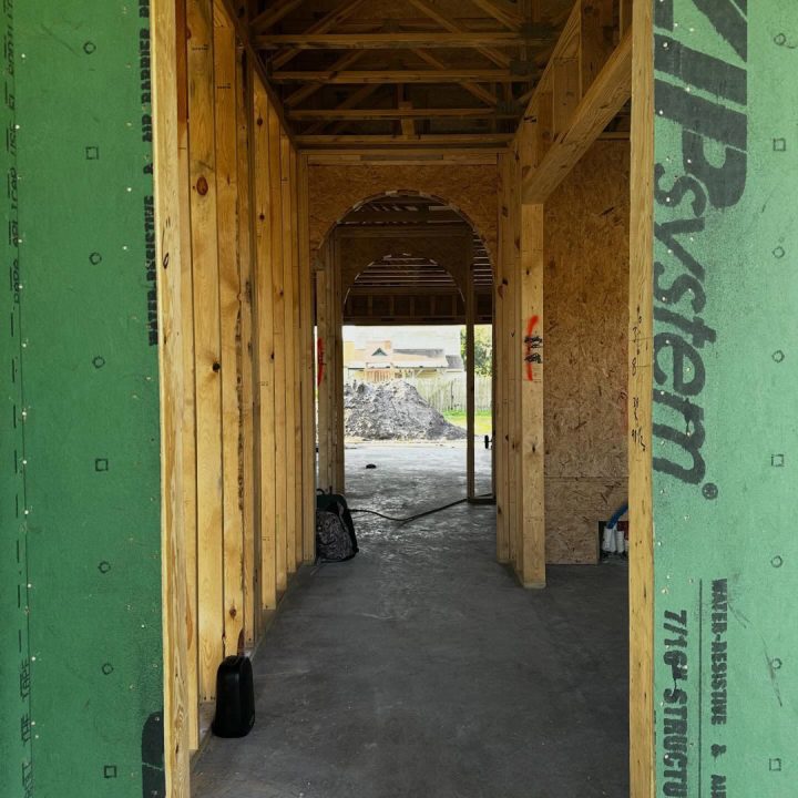 A construction site showing the wooden framework of a building under development, with green sheathing on the exterior walls and an arched doorway opening to the outside.